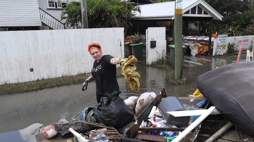 A woman throwing items onto a large pile of rubbish outside her home in low-level flood water.