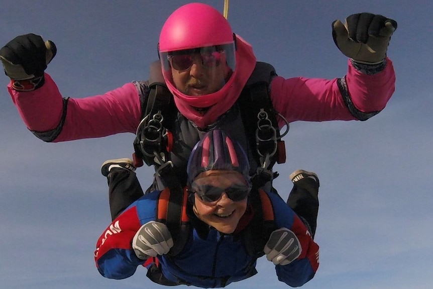 Wendy Mitchell wearing goggles, smiling, flies through the air with a man strapped above, and parachute cord just visible above.
