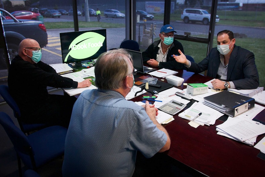 Four men wearing surgical masks talk around a boardroom table.