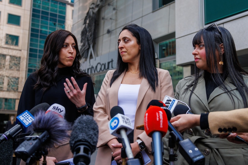 Three women speaking with news microphones in front of them.