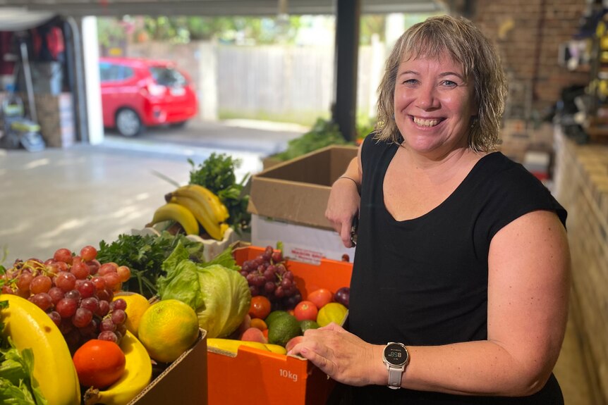 Angela Webster smiles at the camera in front of a box of fruit and veggies which she gets as part of a Sydney food co-op