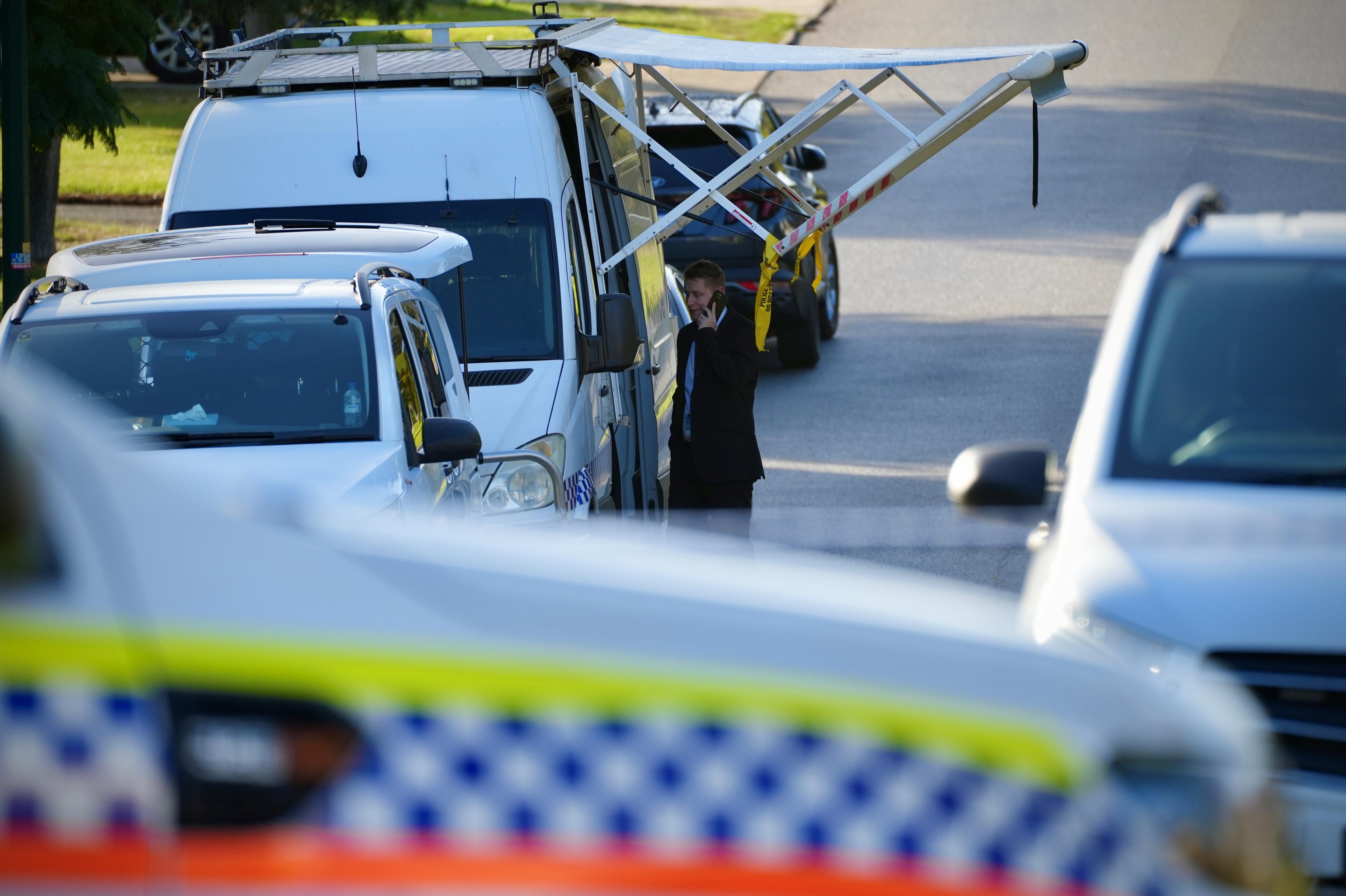 Police and forensic vehicles parked on a suburban street with a man talking on the phone in the middle.