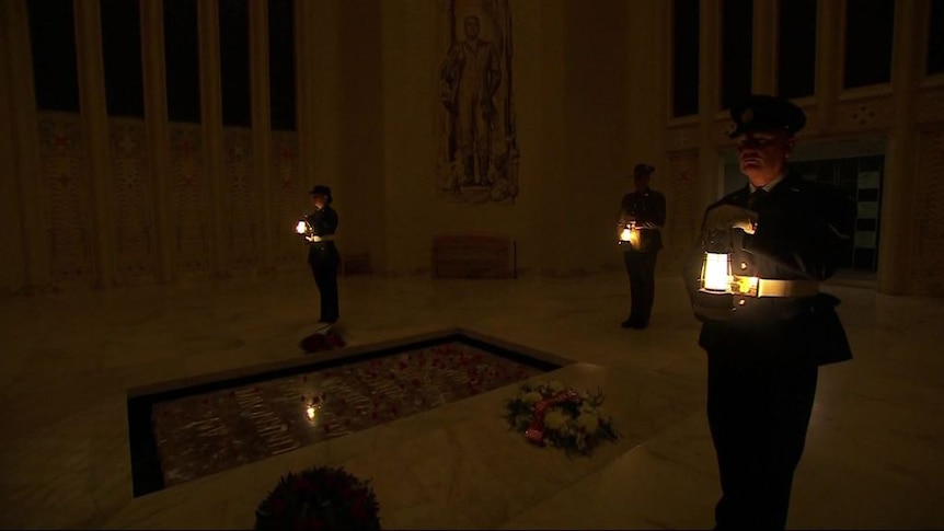 Three soldiers with heads bowed stand in the dim light of dawn holding lanterns at national war memorial.
