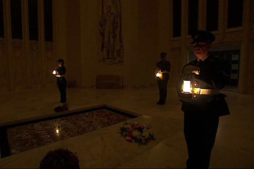Three soldiers with heads bowed stand in the dim light of dawn holding lanterns at national war memorial.