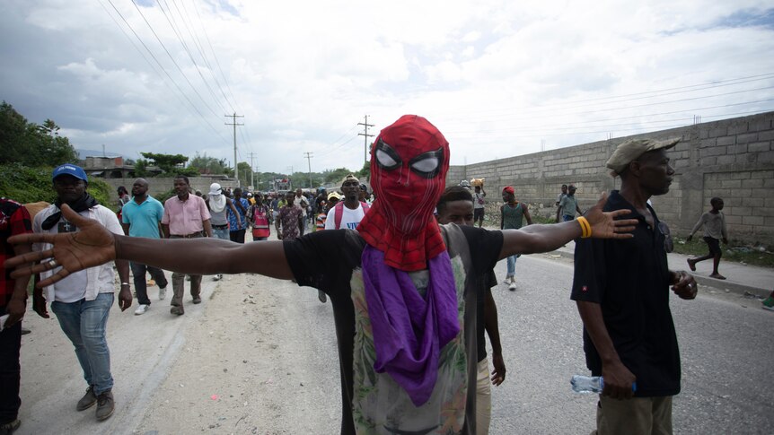 A man wearing a Spiderman mask spreads his arms as he walks down the street with a crowd of people.