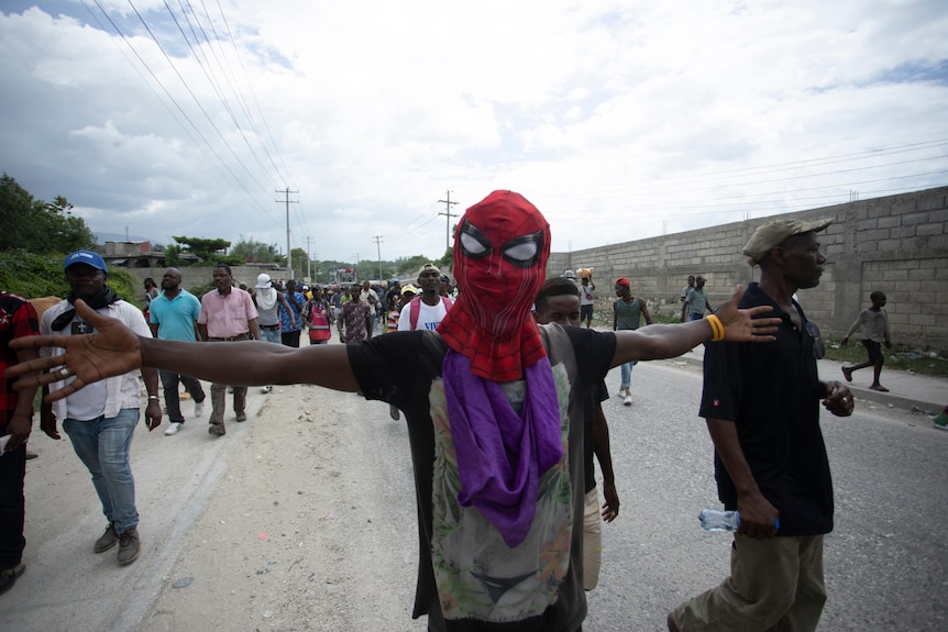 A man wearing a Spiderman mask spreads his arms as he walks down the street with a crowd of people.