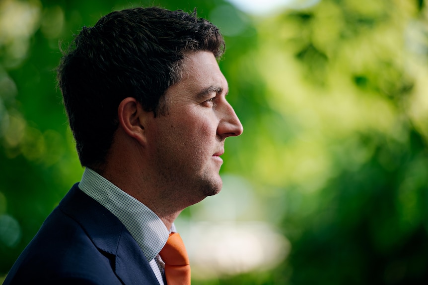 A side-on photo of a man wearing a blue blazer and orange tie. There are blurred out trees in the background.