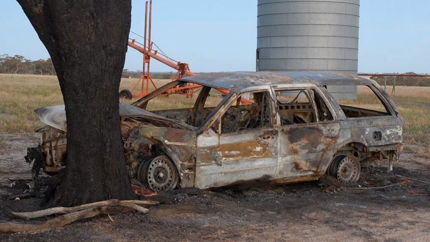 An incinerated car crashed into a tree with farm machinery in the background.