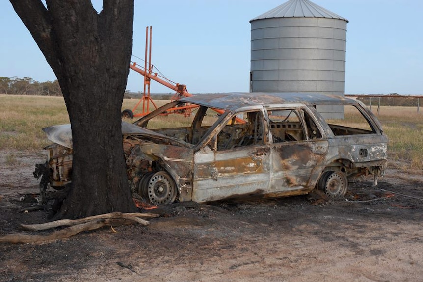 An incinerated car crashed into a tree with farm machinery in the background.
