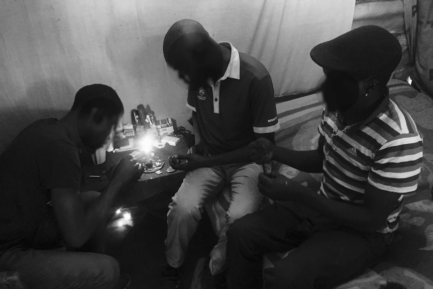 Three men huddle around a table where one is filling a syringe.