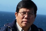 A picture of an older man of Vietnamese heritage with black hair and glasses standing on a boat with the sea in the background.
