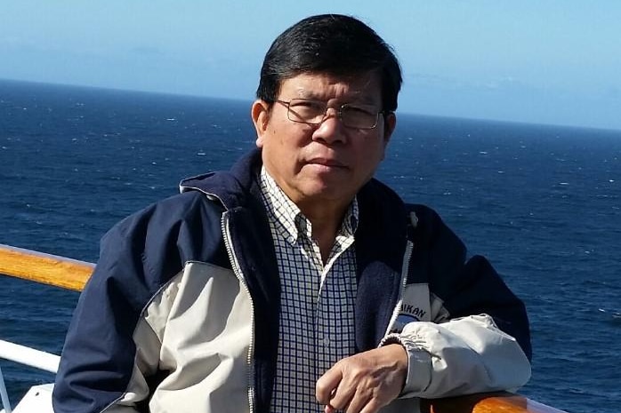 A picture of an older man of Vietnamese heritage with black hair and glasses standing on a boat with the sea in the background.