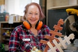 Debi wears a flannelette shirt and orange ear muffs while holding two wooden Christmas trees.
