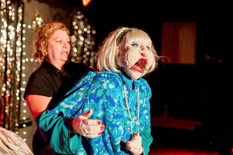 Woman stands behind a life-sized older woman puppet on a stage.