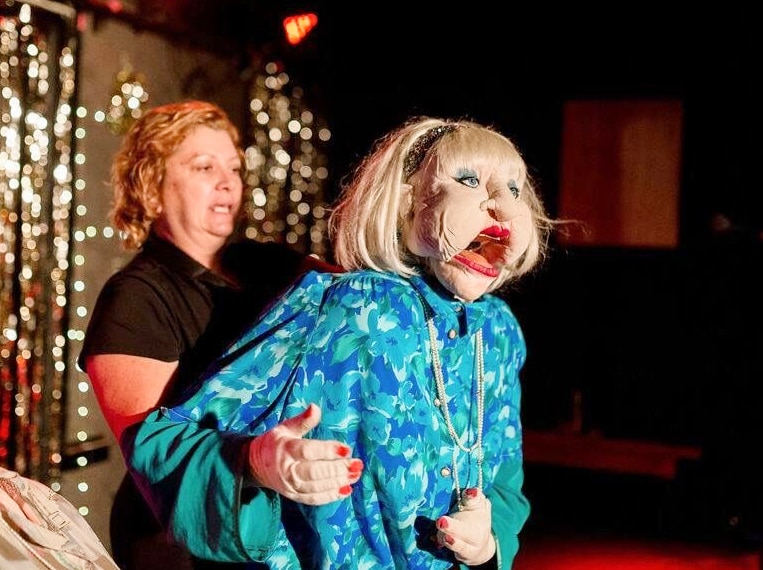 Woman stands behind a life-sized older woman puppet on a stage.