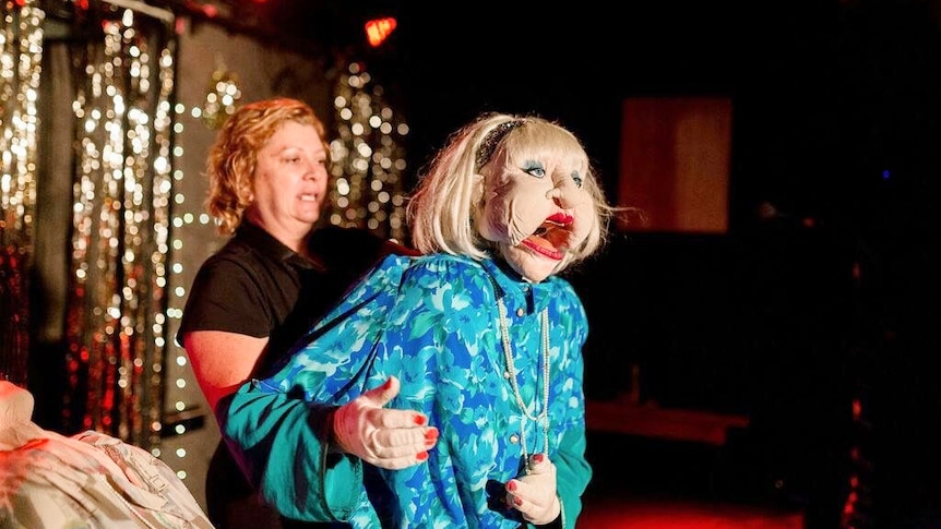 Woman stands behind a life-sized older woman puppet on a stage.