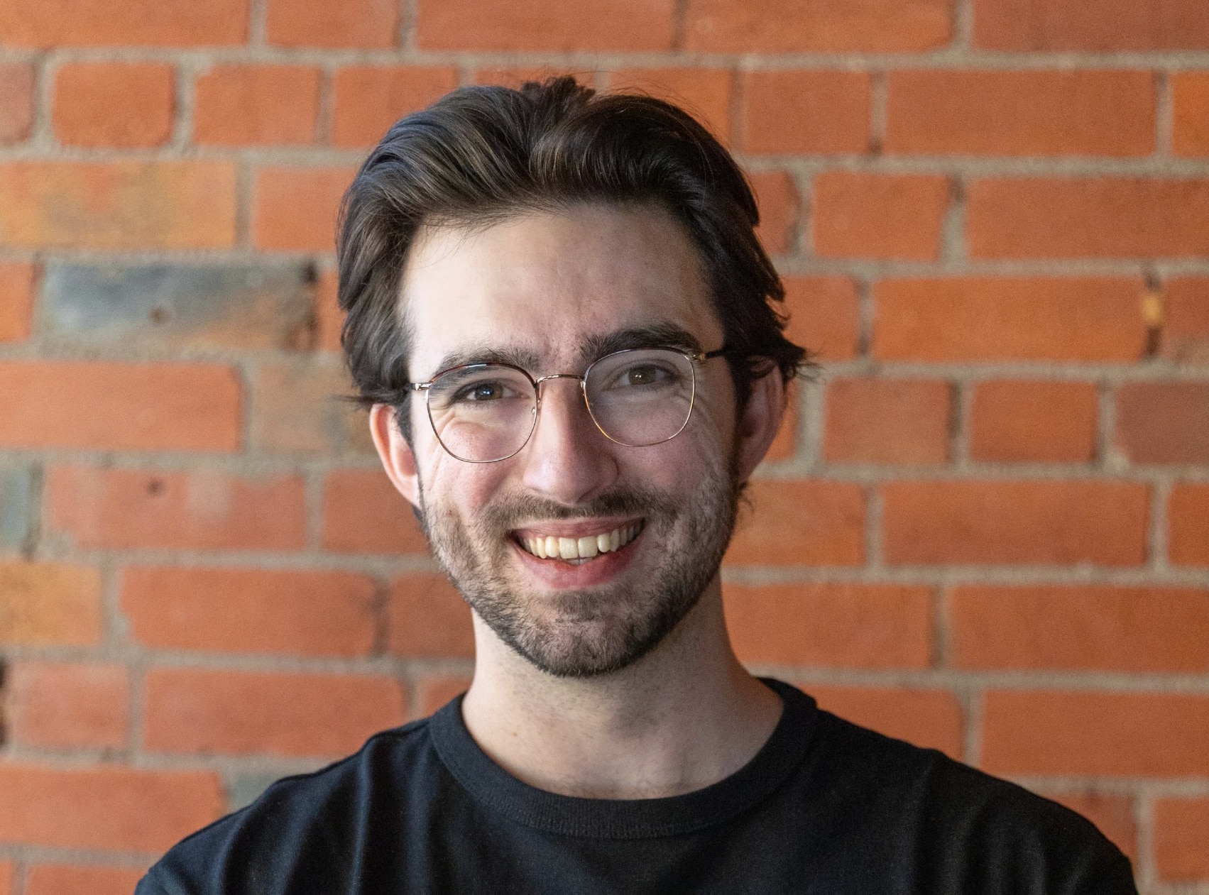 A man wearing glasses and wearing a black shirt stands in front of a brick wall