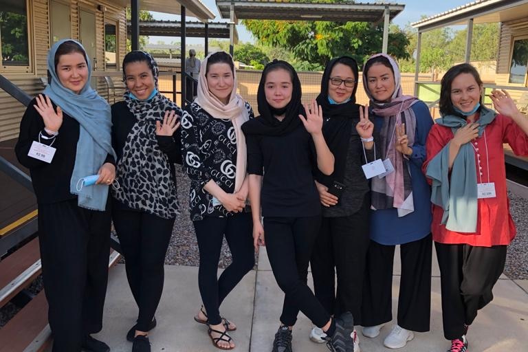 A group of smiling women wave at the camera as they stand next to a building. 