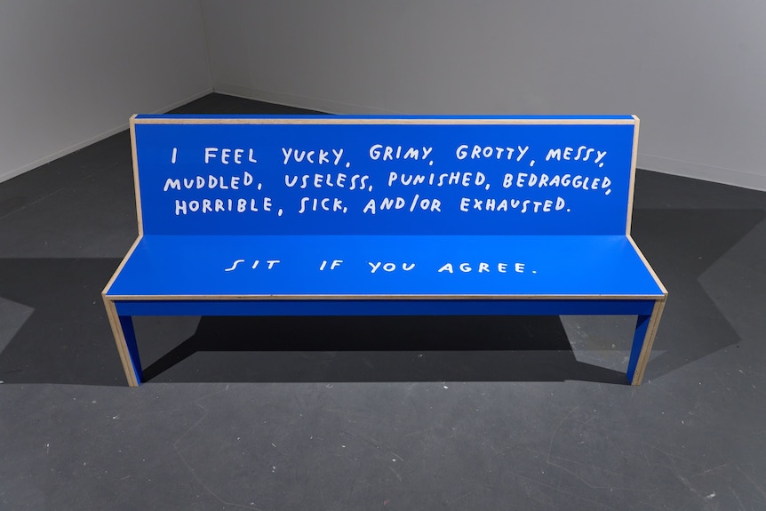 A blue bench on display in a gallery reads: "I feel yucky, grimy, grotty, messy, muddled, useless ... sit if you agree".