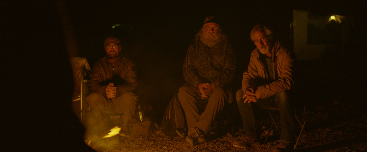 Three men sit on chairs in facing a camp fire, with darkness around them.