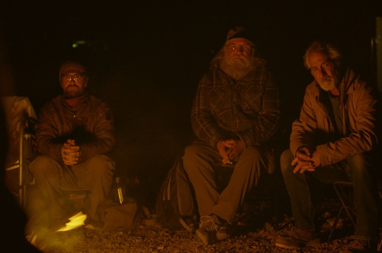 Three men sit on chairs in facing a camp fire, with darkness around them.