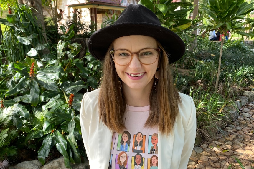 A woman wearing a black hat and with her brown hair down by her side stands in front of some green trees smiling