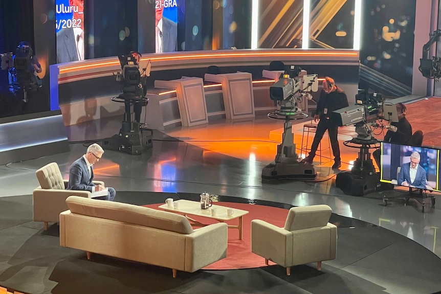 Man sitting on a couch reading notes in a TV studio surrounded by cameras and monitors.