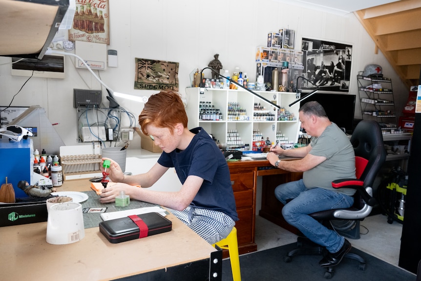 A teenage boy and his father painting scale models in their garage.