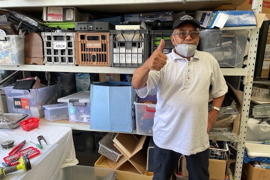 A man gives a thumbs up with second hand goods in shelves behind him. 