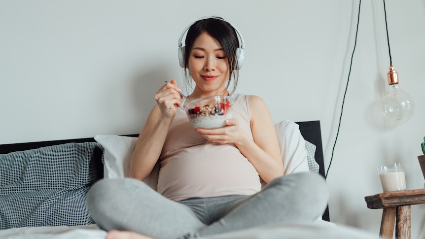 A pregnant woman eating yoghurt with fruit and granola while listening to music with headphones.