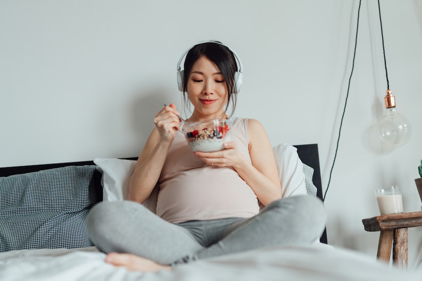 A pregnant woman eating yoghurt with fruit and granola while listening to music with headphones.
