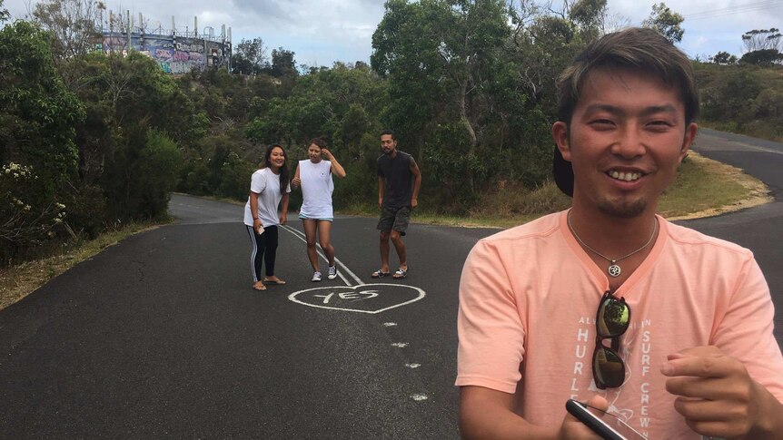 Three Japanese tourists take photos, one of them does the leg splits in the middle of the road, the other sits