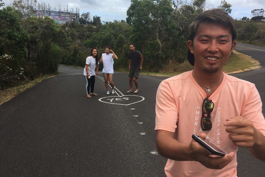 Three Japanese tourists take photos, one of them does the leg splits in the middle of the road, the other sits