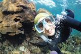 A woman underwater in scuba dive gear with a reef behind her