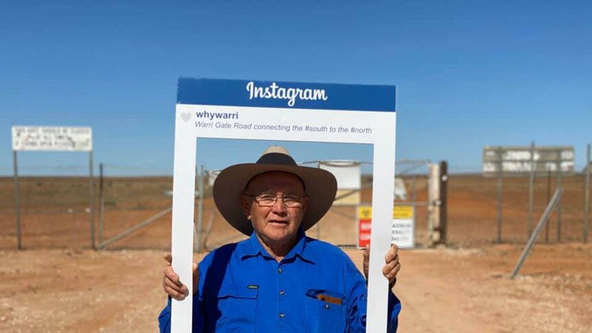 A man in a hat stands on an outback dirt road holding an Instagram frame around his face.