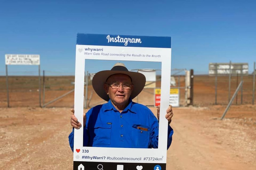 A man in a hat stands with a frame around his face on a dirt road