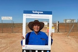 A man in a hat stands on an outback dirt road holding an Instagram frame around his face.