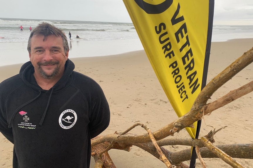Man stands on beach with Veterans Surfing flag behind, he is smiling