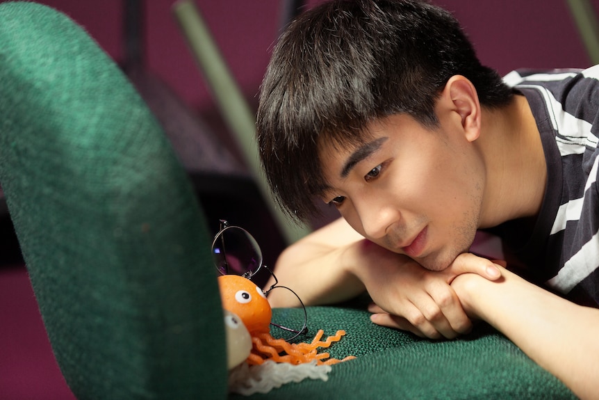 A Chinese man rests his head on his arms on a chair, and looks at glasses and toys on the chair. 