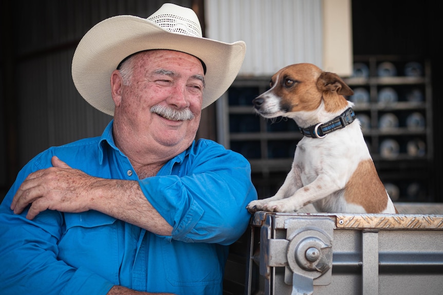 A man wearing a blue shirt, white hat stares at his dog next to him on the back of a ute.