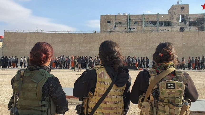 Three women in fatigues watch a group of people outside a prison wall. 
