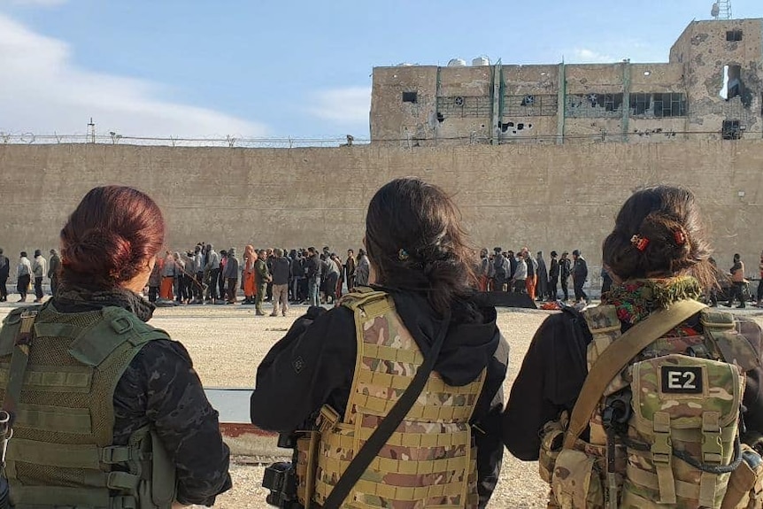 Three women in fatigues watch a group of people outside a prison wall. 