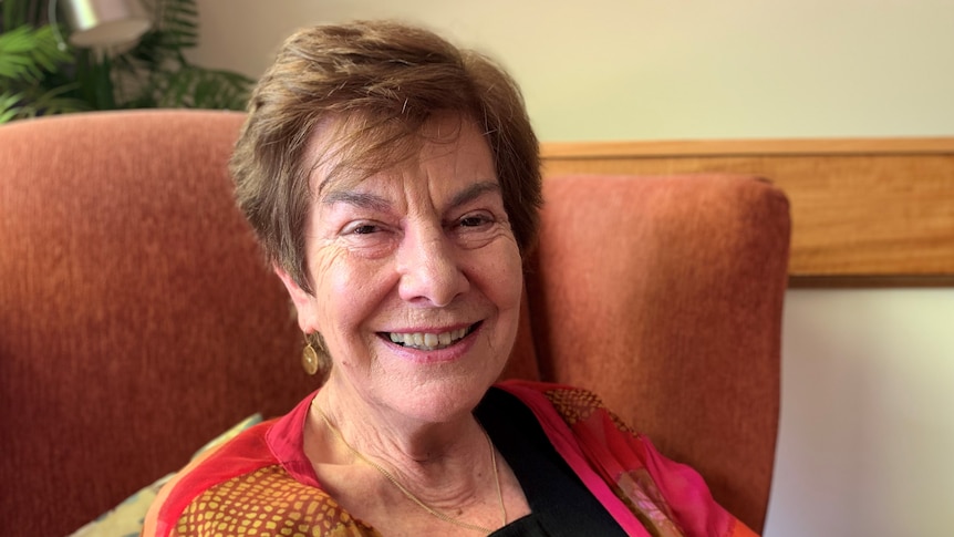 Jennifer Rowe leans back against colourful cushions on a dark orange armchair. She is wearing a bright top and smiling widely