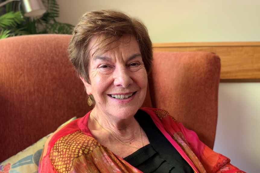 Jennifer Rowe leans back against colourful cushions on a dark orange armchair. She is wearing a bright top and smiling widely