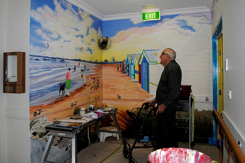 An early man with a walking frame admires a mural of a beach sunset.