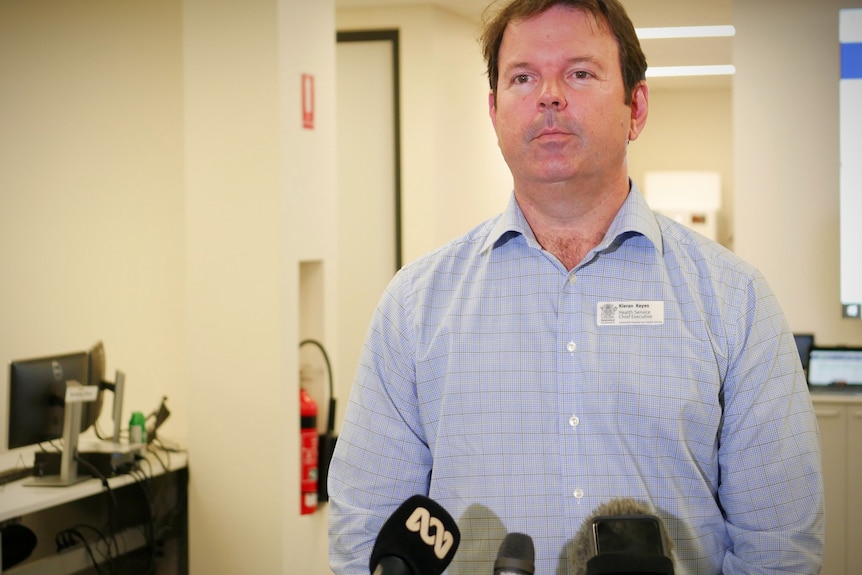 A man in a blue collared shirt stands in an office speaking in front of media microphones