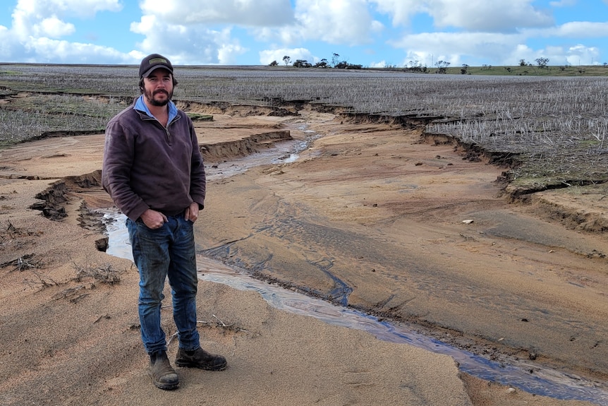 Rain has torn a deep trench in a farmer's paddock. He looks down at the damage.