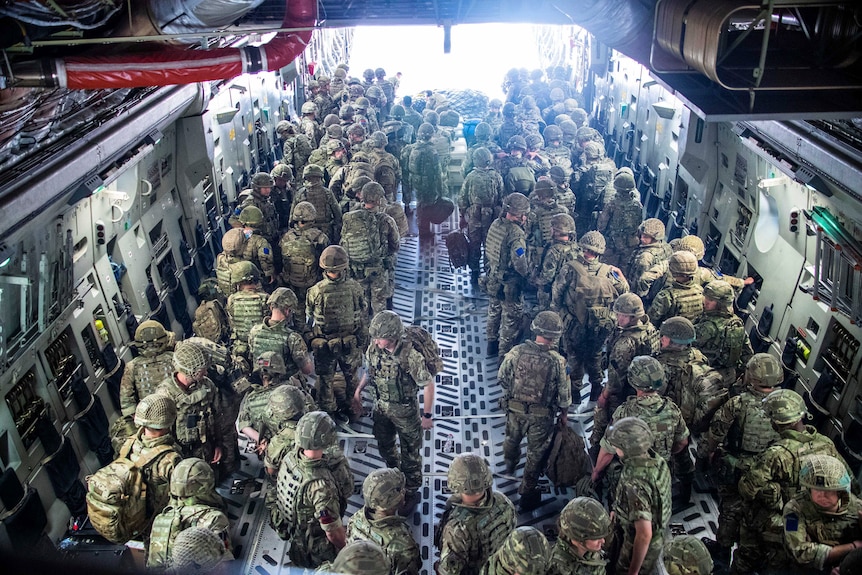 A large group of soldiers in camoflague fatigues and helmets stand ready to walk out of the back of a large aircraft.