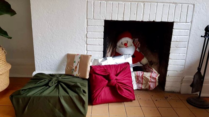 A white fireplace with a Santa doll inside it and fabric-wrapped gifts in red and green on the hearth