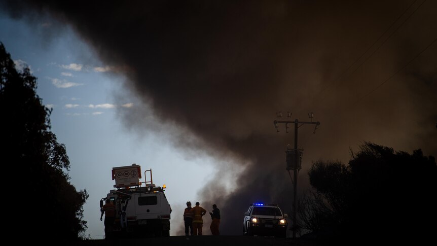 Thick black smoke blankets the sky as two emergency service vehicles respond to a fire.